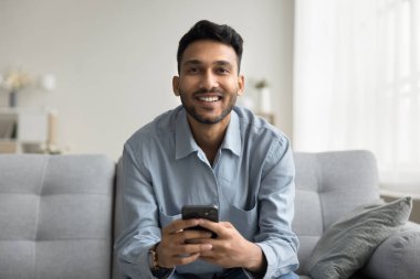 Genç öğrenci işadamı hücresinde kameraya mutlu bir gülümsemeyle bak. İnternetteki basit iletişimle tatmin olmuş bir şekilde oturma odasındaki kanepede oturmuş internet alışverişi şifreleme işini başarıyor.