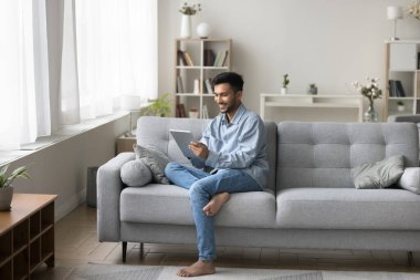 Dijital bir eğlence. Mutlu yakışıklı milenyum erkeği, modern oturma odasındaki kanepede tek başına oturup sohbet oyununda sevgilisiyle mesajlaştığı tablet bilgisayarını tutuyor. İnternetten e-kitap indiriyor. Boşluğu kopyala