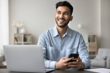 Çoklu görevlerde başarılı. Ev ofisinden bağımsız olarak çalışan mutlu genç işadamı iş yerinde bilgisayar başında otururken akıllı telefonu elinde tut kendine güvenen bir gülümsemeyle aradıktan sonra kenara bak.