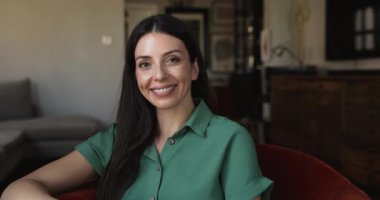 Modern oturma odasındaki rahat koltuğa oturmuş kameraya poz veren yeşil gömlekli güzel bir kadın, hafta sonları kendi evinde ya da kiralık evinde dinleniyor. Refah, ev sahibi portresi
