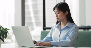 Dizüstü bilgisayarda çalışan Asyalı kadın, görevlere odaklanmış, belge yazma veya düzenleme, düşünme, araştırma yapma veya verileri analiz etme, e-postalara yanıt verme, modern çalışma alanında sunum hazırlama veya rapor hazırlama