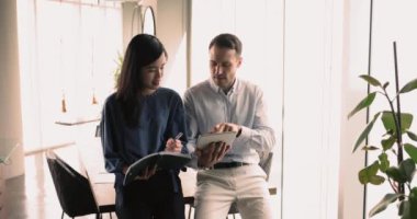 İş adamı genç Asyalı çalışan kadını eğitiyor, tablet üzerinde online içeriği gösteriyor ve açıklıyor, şirket desteği veriyor, genç meslektaşlarına notlar yazma tavsiyesi veriyor.