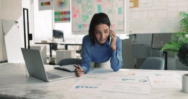 Profesyonel kadın, proje yöneticisi veya finansal analist dizüstü bilgisayarla çalışıyor, grafiklerle, istatistiksel bilgilerle belgeyi inceliyor, iş arkadaşı veya müşteriyle telefonda konuşuyor