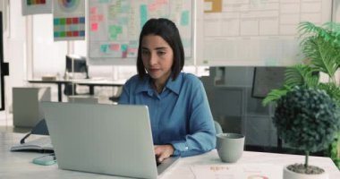 Modern ofiste masa başında oturan genç profesyonel kadın dizüstü bilgisayarını kullanarak grafikler ve diyagramlar ile basılı belgelere dikkat çekiyor. Veri analizi, raporlama, stratejik planlama