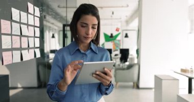 Dijital tablet üzerinde çalışan profesyonel iş kadını, modern cihaz hakkındaki bilgileri gözden geçiriyor, verileri planlıyor, analiz ediyor veya görevleri yönetiyor, ortak çalışma odasındaki yapışkan notların yanında duruyor. Çalışma Akışı