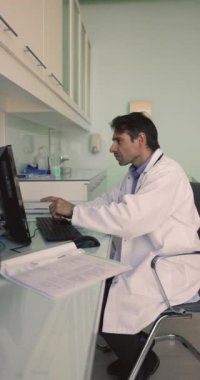 İşyerinde bilgisayar kullanan orta yaşlı bir tıp uzmanı, hastaların verilerini kağıttan sisteme taşıyor, klavyede yazıyor, belge okuyor, monitöre işaret ediyor, dikey çekim yapıyor.