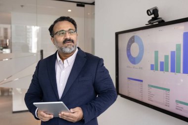 Düşünceli yetişkin Hintli iş adamı iş projesi stratejisi üzerinde çalışıyor, sunum odasında, elinde tablet bilgisayarla, elinde pazarlama raporuyla büyük TV ekranında duruyor.