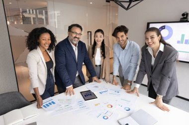 Çok ırklı genç meslektaşlar ve yaşça büyük liderlerden oluşan bir ekip, büyük toplantı masasında birlikte duran patron, kameraya bakıyor, gülümsüyor, grup fotoğrafı için poz veriyor.