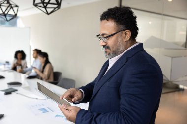 Yetişkin Hintli iş adamının toplantı masasında dijital tablet kullanması ve arka planda çalışanlardan oluşan farklı bir ekibin olması. Orta yaşlı işletme koçu eğitim seminerine hazırlanıyor.