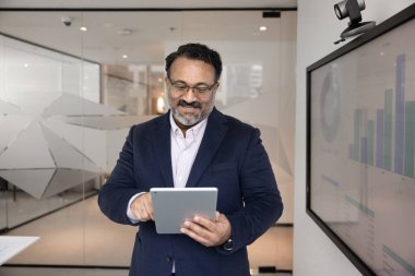 Olgun yetişkin Hintli işadamı sunum odasında tek başına çalışıyor, elektronik kurulda duruyor, tablet pazarlama raporunu analiz ediyor, dijital aletleri iş iletişimi için kullanıyor, gülümsüyor.