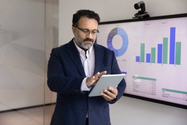Ciddi Hintli kıdemli iş adamı, pazarlama verilerini tablet bilgisayarında analiz ediyor, dijital cihazlarla daktilo yazıyor. İş öğretmeni büyük TV ekranında sunum hazırlıyor