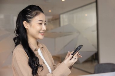 Neşeli başarılı genç Asyalı satış müdürü iş yerinde cep telefonunu kullanıyor, cep telefonuyla yazıyor, internetten sohbet ediyor, mesaj atıyor, gülümsüyor, gülüyor, çevrimiçi iletişimden keyif alıyor.