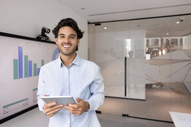 Kendine güveni tam, hırslı genç bir iş adamı elinde tablet, kameraya bakıyor, gülümsüyor, ofiste eğitim odasında pazarlama verileriyle poz veriyor.