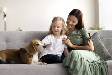 Sevimli anaokulu öğrencisi küçük kızı ve annesi kanepede oturuyor, nazikçe köpeğini okşuyor, sıcak ve sevgi saçıyorlar. Aile bağları, güven ve arkadaşlık, evcil hayvanlarla insanlar arasındaki duygusal bağ.