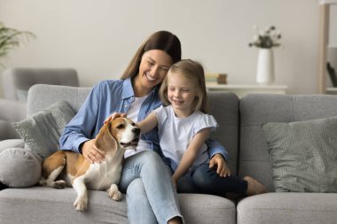 Sevimli kız rahat koltukta oturan sevimli av köpeğini okşuyor. Sevgi dolu genç annesiyle birlikte. Aile, evde kaliteli vakit geçiriyor.