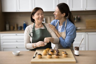İki farklı nesil kadın, genç yetişkin kız olgun gülümseyen annesine sarılıyor, kahkahalarla gülüyor modern mutfakta pişmiş kek dekorasyonunun keyfini çıkarıyorlar, sıcaklık, neşe ve sevgi saçıyorlar. Aile yemeği zamanı.