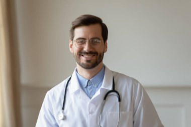 Gözlüklü ve steteskoplu beyaz önlüklü gülümseyen genç doktor portresi kameraya bakıyor, dost canlısı pratisyen hekim ofiste fotoğraf için poz veriyor, sağlık konsepti...