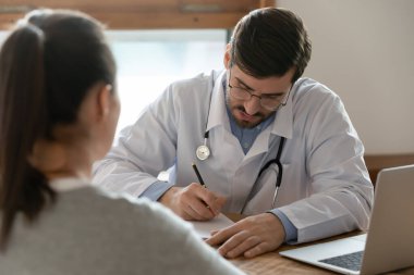 Hasta kartına reçete yazan odaklanmış bir doktor kadınla tıbbi randevuda, anestezi, hastalık geçmişi, sağlık ve sigorta kavramını dolduran ciddi bir erkek GP terapisti.