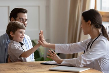 Gülümseyen küçük çocuk, beyaz üniformalı kadın doktora beşlik çakıyor, hastanede baba kucağına oturuyor, çocuk doktoru, okul öncesi hastayı babasıyla selamlıyor.