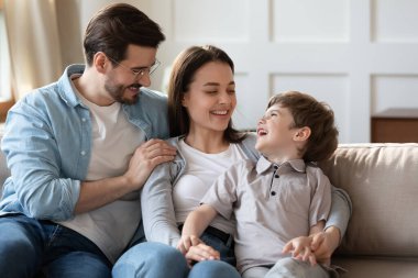 Küçük oğluyla mutlu bir aile, rahat bir koltukta oturup gülüyor, baba gözlük takıyor ve anne anaokulu çocuğuyla eğleniyor, hafta sonları evde boş vakitlerin tadını çıkarıyor.