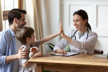 Gülümseyen kadın doktor, steteskopla beyaz üniforma giymiş, babasının muayenesinde küçük bir hastayı karşılıyor, anaokulu çocuğu babasının kucağına oturuyor, pediyatriste beşlik çakıyor.
