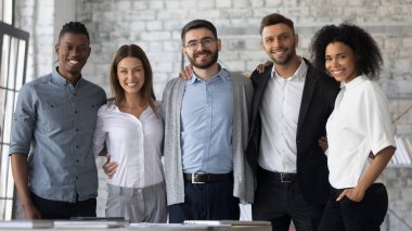 Çeşitli etnik çeşitlilikte gülümseyen genç iş adamlarının grup portresi ofiste poz veriyor, iş yerinde birlik ve destek gösteriyor, mutlu motivasyonlu çok ırklı meslektaşlar takım ruhunu gösteriyor