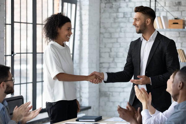 Happy caucasian businessman shake hand greeting with promotion excited african American female employee at meeting, smiling male boss handshake get acquainted with biracial woman worker at briefing