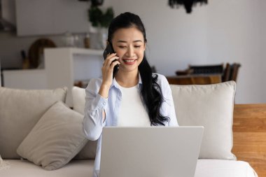 Olumlu meşgul genç Asyalı kadın evden çalışıyor, cep telefonuyla konuşuyor, gülümsüyor, gülüyor, sohbet sırasında dizüstü bilgisayar kullanıyor, kanepede oturuyor, iletişimin tadını çıkarıyor.