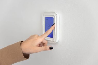 Duvara monte edilmiş akıllı ev aleti kullanan, termostatı programlayan, ekrandaki hava sıcaklığını düzenleyen, klimayı başlatan, cihazdaki düğmeye basan bir kadın eli. Yakın çekim kesilmiş.
