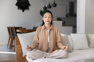 Sakin, genç Asyalı bir kadın kanepesinde meditasyon yapıyor, sukhasana pozu veriyor, zen parmaklar takıyor, derin meditasyonda kanepede oturuyor, temiz hava soluyor, dikkat çekiyor.