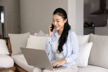 Cep telefonuyla konuşan, cep telefonu kullanırken dizüstü bilgisayar kullanan, evden internet projesinde çalışan, rahat koltukta bilgisayarda yazan mutlu, genç Asyalı serbest yazar kız.