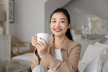 Mutlu, rahat, genç Asyalı bir kadın sabah kahvesini yudumlarken elinde seramik bardaklar, sıcak bir içecek, rahat bir kanepeye oturmuş, kameraya bakıyor, portre için gülümsüyor.