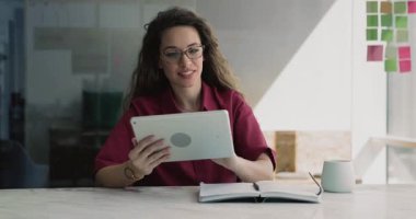 Gözlüklü genç bir kadın iş yerinde tablet üzerinde çalışıyor, gülümsüyor, uzaktan etkileşimden hoşlanıyor, çevrimiçi proje için araştırma bilgileri, dijital araçlar veya iş için yapay zeka platformu kullanıyor