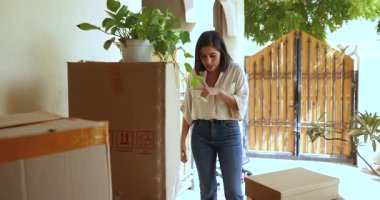 Ön bahçede, terasta karton lojistik kutuları kontrol eden ve taşıyan genç Latin bir kadın var. Mutlu yeni kiracı ya da ev sahibi daireye taşınıyor