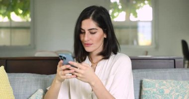 Odaklanmış genç, güzel İspanyol bir kadın evde akıllı telefon kullanıyor, cep telefonuyla yazı yazıyor, heyecanlanıyor, mutlu oluyor, nefes nefese kalıyor, gülüyor, zafer kazanıyor.