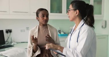 Genç Afrikalı kadın hasta sağlık sorunlarını paylaşıyor, elinde dosya tutmakta olan Avrupalı kadın pratisyen doktorla konuşuyor, şikayetler, semptomlar, potansiyel teşhisler, testler veya prosedürleri tartışıyor.