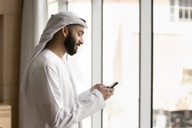 Yan görünüm gülümseyen Arap işadamı geleneksel beyaz bulaşık makinesi elbisesi giyiyor modern akıllı telefonu tutuyor, arıyor, okuyor, e-postaları kontrol ediyor, mesajlara cevap veriyor, yeni cep telefonu uygulamaları indiriyor.