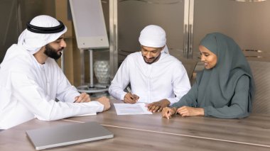 Arap bir çift ve bir yönetici konferans masasında oturuyor. Geleneksel kandora gibi giyinmiş bir adam yasal belgeleri imzalıyor. Resmi bir anlaşmaya imza atıyor.