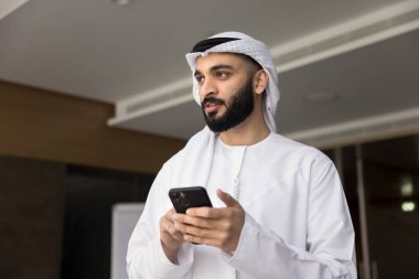 Genç Arap iş adamı geleneksel Ortadoğu bulaşık giysisi içinde elinde akıllı bir telefonla ofiste duruyor, modern teknolojiyi kullanarak iş akışı sağlıyor.