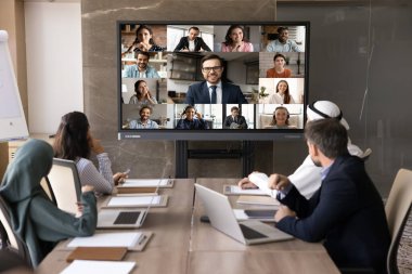 Çok kültürlü iş adamlarından oluşan bir grup video konferansına katılıyor, konferans masasında oturup çeşitli iş ortaklarını gösteren TV ekranlarına bakıyorlar, sanal bir toplantıya giriyorlar, uzaktan pazarlık yapıyorlar.