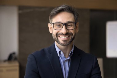 Resmi takım elbise giymiş yakışıklı Latin işadamı modern ofiste gözlük takıyor, kameraya neşeli bir ifadeyle bakıyor, kariyerinden tatmin oluyor, yetkinlik ve profesyonellik yayıyor.