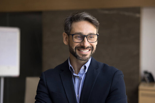 Handsome Portuguese middle-aged businessman in formal jacket and glasses posing in office, looking away. Head shot portrait of company owner, CEO or executive manager feel satisfied with achievements