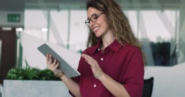 Profesyonel iş kadını, modern iş aletleri ve platformları kullanarak tablet üzerinde çalışan gözlük takar, çağdaş kurumsal çalışma alanında durur, yeni görevleri gözden geçirir, rapor hazırlar