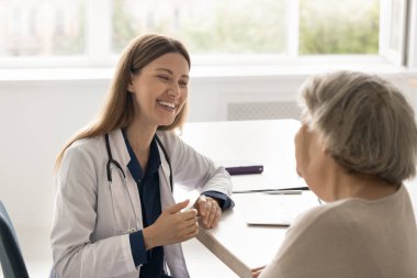 Sağlık hizmeti sohbeti. İçten gülümseyen genç bir kadın doktor. Klinikteki masasına oturup yaşlı kadın hastalarla iletişim kurarak yardım etmek isteyen semptomları dinle.