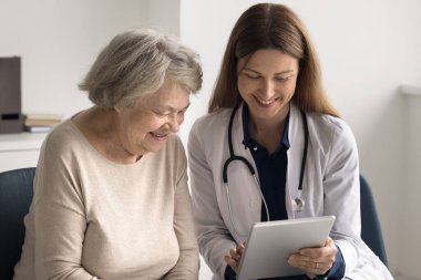 Sağlık sektöründe bilgisayar yenilikleri. Gülümseyen dost canlısı genç bayan doktor aile terapisti yaşlı kadın hastaya dijital tablet ekranı göster. Yaşlılara sağlık uygulamasının nasıl kullanılacağını öğret.