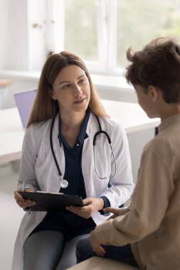 Doktor randevusunda. Nazik, genç bir bayan çocuk doktoru aile terapisti klinikteki küçük hasta çocukla röportaj yapıyor sağlık durumuyla ilgili sorular soruyor. Kağıt tıp dosyasına not alın.