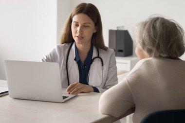 Tıpta modern teknoloji. Genç bayan doktor aile terapisti klinikteki masada oturuyor yaşlı bayanla sağlık kontrolü için buluşuyor hastalık hakkında bilgi vermek için not defteri kullanıyor elektronik hasta kayıtlarını okuyor.