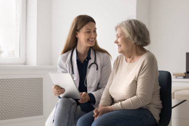 Sonuçların iyi. Gülümseyen dost canlısı genç doktor yaşlı kadını muayene ediyor. Tablet bilgisayarındaki hasta dosyalarına kayıt tutuyor. Önceki testlere kıyasla pozitif dinamik görüyorlar.