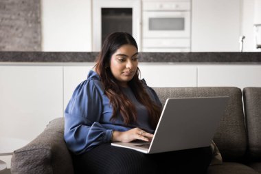 Genç Hintli kadın kanepeye oturmuş, bilgisayarında çalışıyor ya da geziniyor modern ve rahat oturma odasında tek başına dinleniyor. Eğitim platformunu kullanarak iletişim, araştırma, uzaktan çalışma