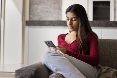 Ciddi Hintli genç bir kadın rahat bir koltukta modern akıllı telefonuyla dinleniyor, kısa mesajlar gönderiyor veya okuyor, telefon görüşmeleri yapıyor veya alıyor, web sörfü bilgileri alıyor, evde boş zamanlarını internette geçiriyor.